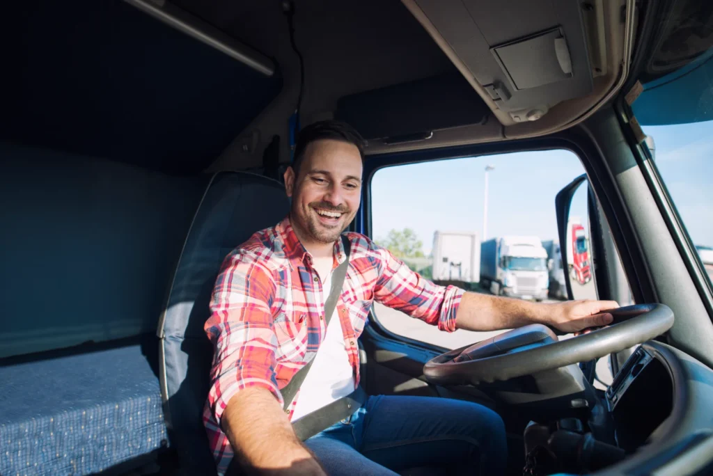 Man in plaid shirt driving a truck, smiling at the wheel inside the cab.